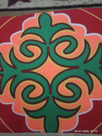 The image shows a close-up view of a red fabric with a green, orange, and yellow stylized floral or ornamental pattern. The pattern is outlined in dark red with a jagged texture. There is a timestamp in the bottom right corner: "05.07.2026 07:13". The image is a photograph of a textile, possibly a cushion or part of a larger piece of fabric, placed on a surface that appears to be carpeted with a brown and cream patterned rug in the background. The location is identified as Жаңақұрылыс, Kazakhstan.