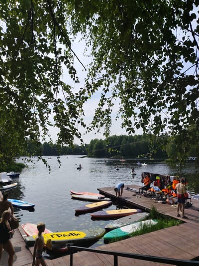 This image depicts a recreational waterfront scene at a lake or river, likely in or near Vylytydor, Russia, given the Cyrillic text on a paddleboard that reads "ГРААН" (GRAAN). In the foreground, a wooden dock is visible, with several paddleboards and a young boy standing on it. Further back, a woman and a man are walking on the dock. The water is dotted with several people on paddleboards and kayaks, engaged in various activities. A rack with life jackets is also present on the dock. The background features a dense line of green trees under a cloudy sky. The overall atmosphere suggests a summer day at a watersports facility.
