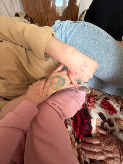 In the city of Valozhyn, Belarus, two individuals, one adult and one child, are seen from the chest down, sitting close together. The adult, wearing a tan jacket and blue jeans, has the word "BELA" written in pink marker on their hand. The child, dressed in a pink sweater, has a drawing of a heart with dots and leaves on their hand. Both individuals have their arms intertwined, with the child's arm resting on the adult's chest. They are seated on a floral-patterned blanket. The background suggests an indoor setting with a wooden partition visible.