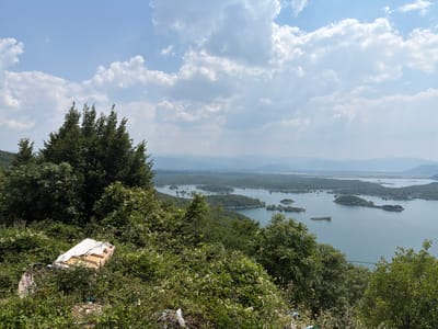 An aerial, wide-angle shot captures a tranquil, expansive lake dotted with numerous tree-covered islands under a bright, partly cloudy sky. The lake stretches towards a hazy, mountainous horizon. In the foreground, the dense foliage of trees and bushes partially obscures a discarded wooden frame covered with a white sheet and scattered litter. The scene is situated in the vicinity of Ponikvica, Montenegro.