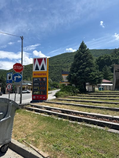 The image, taken in Vetren Dol, Bulgaria, depicts a sunny daytime scene featuring a roadside gas station against a mountainous backdrop. In the foreground, a gray wheeled trash can is visible on the right. A utility pole with power lines extends from the left.
Dominating the midground is a tall yellow and red gas station sign, displaying the name "МЕЧКАРОВ" (MECHKAROV) in Cyrillic and a distinctive "M" logo. Current fuel prices are listed: LPG at 0.75, DIESEL at 1.71, A95H at 1.56, and A100 at 6. The sign also indicates available services with icons for WC, WiFi, and acceptance of VISA cards. Behind the sign, a white car is partially visible.
To the left of the gas station sign, several road signs are mounted on a pole: a red octagonal "STOP" sign, a blue circular sign indicating a right turn, a blue rectangular sign showing a gas pump, and a blue circular sign with the number "5".
On the right side of the image, a series of concrete steps with grass growing between them ascends a slope. Further back, buildings with red roofs are nestled among trees. The background features lush green mountains under a bright blue sky dotted with scattered white clouds, suggesting clear and pleasant weather. The overall scene is one of a functional roadside stop integrated into a natural, scenic environment.