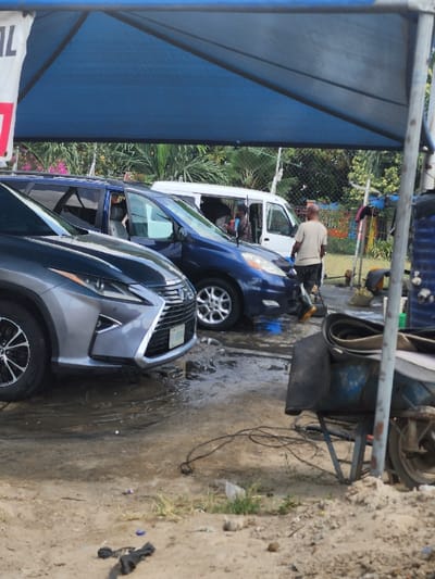This image depicts an outdoor car wash or vehicle service area in Igando Oloja, Nigeria. The scene is dominated by a large, blue tarpaulin canopy providing shade over multiple vehicles.

In the foreground, a silver/grey Lexus SUV is parked on the left, displaying a Nigerian license plate. To its right, a dark blue minivan or SUV is actively being washed; a male individual, dressed in a tan short-sleeved shirt and dark shorts, stands beside it, holding a yellow object consistent with a sponge or brush. Water and muddy puddles are visible on the packed dirt and gravel ground beneath and around the vehicles. Further back and to the right of the blue minivan, a white van is parked, with a person partially visible near its open door. Another individual is partially visible behind the blue minivan near a blue barrel-like object.

The ground surface is uneven, consisting of dirt and gravel, with areas of standing water and mud. Scattered black cables or hoses lie on the ground in the mid-foreground. To the far right, a blue metal wheelbarrow containing dark, possibly rubber, objects is positioned near a canopy support pole. In the background, lush green tropical foliage and what appears to be a chain-link fence are visible under clear skies. A white banner with the partial text "AL" (in red) hangs from the canopy structure on the far left.