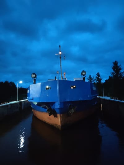 A blue and tan barge is positioned within a concrete lock structure at dusk. The sky above is a deep blue with visible clouds. The barge has a prominent blue upper structure with two circular lights mounted on poles. The lower hull of the barge is tan. The water in the lock reflects the lights and the sky. Trees and a lamppost are visible along the sides of the lock. The scene appears to be in the city of Devyatiny, Russia, given the context provided.