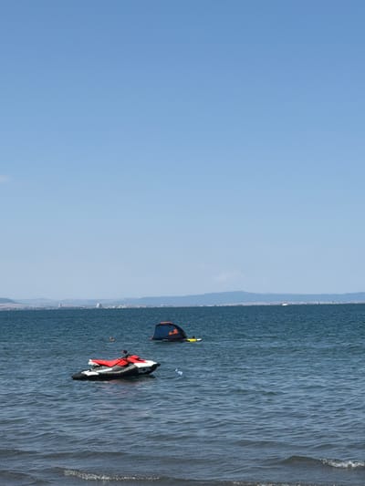 The image depicts a wide, calm body of water under a clear blue sky, with a distant, hazy shoreline. In the foreground, the water exhibits gentle ripples. Two objects are visible on the water. To the left, a red and white jet ski is floating, seemingly stationary. To the right, a dark blue and orange tent-like structure is partially submerged, with a person in yellow attire visible near it. The distant shoreline features a line of buildings, suggesting a coastal town or city, possibly Atiya, Bulgaria. The overall impression is one of a serene day at sea.