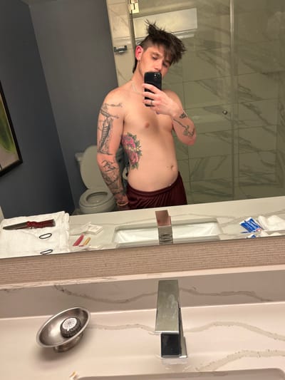 The scene depicts a male individual taking a mirror selfie in a modern hotel bathroom located in Reno, United States of America. The subject is shirtless, presenting a dark, spiky haircut with shaved sides and a longer top, consistent with a modern mullet. He displays extensive tattoos, including a full black and grey sleeve on his left arm featuring figures and intricate designs, and a large colored rose tattoo with green leaves on his left rib cage. Additional tattoos are visible on his right arm. He wears dark red athletic shorts or underwear, a thin silver chain necklace, a silver ring on his left ring finger, and a light-colored beaded bracelet on his right wrist. He holds a dark-colored smartphone, consistent with an iPhone, in his right hand, angled to capture his reflection.

The bathroom features a white vanity countertop with grey veining, a rectangular undermount sink, and a polished chrome single-lever faucet. The wall behind the subject is painted a dark blue-grey. To the immediate left, a white toilet with an open lid is partially visible in the reflection. In the reflection to the subject's right, a modern shower stall with clear glass panels and white tiled walls is apparent.

On the left side of the vanity countertop, a white folded towel supports a red-handled folding knife with a silver blade and two black hair ties. Near the sink, a small silver oval dish contains a circular object, possibly a decorative bottle cap or patterned soap, embossed with a radial design. Further to the right on the countertop, several small packaged items, including red and yellow packets, are visible. Additional white towels and dental hygiene products, such as a toothpaste tube and toothbrush, are reflected on the far right side of the counter.