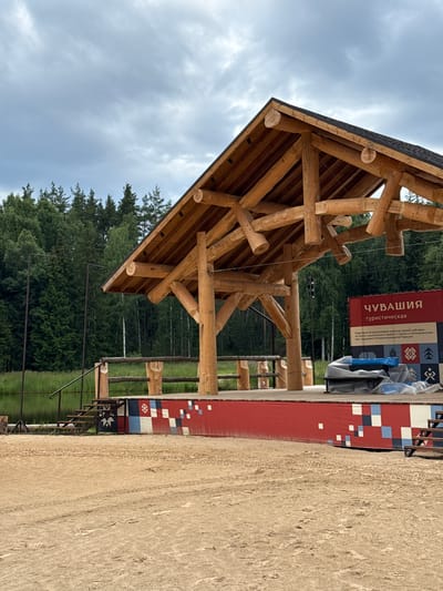 The image shows a wooden log structure with a gabled roof, likely a pavilion or shelter, set in a natural outdoor environment. The structure is made of large, raw-hewn logs, giving it a rustic appearance. Beneath the roof, a complex arrangement of wooden beams and supports is visible.

In the background, a dense forest of green trees stretches out under a cloudy sky. The sky itself is overcast, suggesting a potentially cool or rainy day. To the right of the structure, there is a sandy area, possibly a path or playground, and a red wall adorned with a mosaic of colored squares in red, white, blue, and black.

A banner on the red wall displays Russian text. The visible text reads: "ЧУВАШИЯ туристическая" (Chuvashia Tourist). Below this, there is more text which appears to be descriptive of an event or initiative related to tourism in the Chuvashia region, mentioning a national project and support from the Ministry of Economic Development of Chuvashia.

The scene appears to be an outdoor recreational area or a tourist information point within a natural setting. There are no people visible in the image, focusing the attention on the architecture and the surrounding environment. The overall impression is one of a newly constructed or developing tourist facility in a forested, natural landscape.