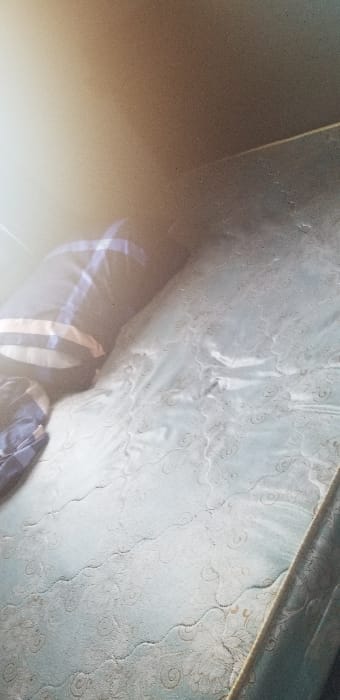 This image depicts an unmade bed, likely located within a private residence in Ado-Ekiti, Nigeria. The central subject is a light blue or greenish-blue mattress with a white or off-white quilted floral and swirling pattern. The mattress shows signs of wear and surface dust or debris. A dark blue pillow with white and light blue horizontal stripes is positioned on the upper left portion of the mattress. Part of a crumpled dark blue and white plaid fabric, possibly a blanket or sheet, is visible below the pillow. The background on the upper left is an overexposed area of light-colored wall or ceiling, indicative of ambient light source or bright reflections. The overall environment appears to be an interior space, likely a bedroom, with no individuals present. No specific actions or interactions are depicted.