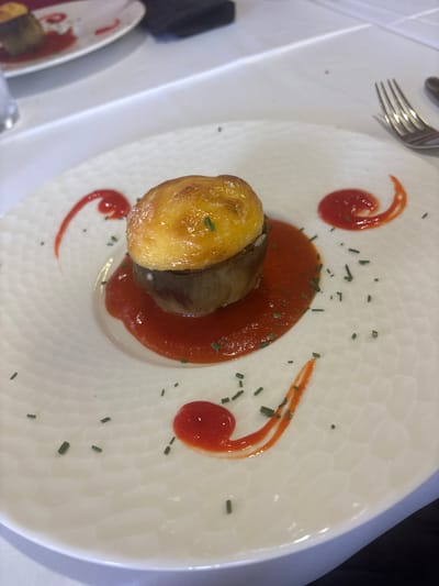 A close-up, eye-level shot captures a single serving of stuffed eggplant on a white plate. The eggplant, a cylindrical piece with a golden-brown, pastry-like topping, rests in a pool of vibrant red sauce. The plate itself features a subtle, textured pattern. Red sauce is artfully swirled onto the plate in decorative patterns, and finely chopped chives are sprinkled around the dish. The setting appears to be a restaurant in Urbanización Roquetas De Mar, Spain, suggested by the white tablecloth and the presence of a fork on the right side of the frame, indicating a dining environment. The lighting is bright, focusing attention on the food presentation.