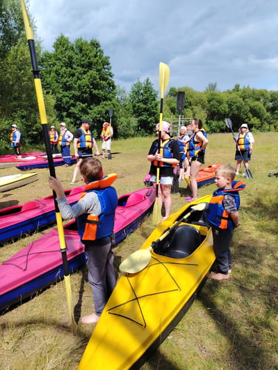A group of people are gathered in a grassy field with several kayaks and paddles. The subjects include a young boy in the foreground wearing an orange and blue life jacket, holding a yellow and black paddle. Behind him, a group of adults and some older children are also wearing life jackets and holding paddles. Some of the kayaks are pink and blue, while one in the foreground is bright yellow. The scene appears to be outdoors, likely a park or recreational area, with trees and a cloudy sky visible in the background. The overall atmosphere seems relaxed and preparatory, suggesting an upcoming kayaking activity. The time of day appears to be daytime, given the lighting. No text is visible in the image.