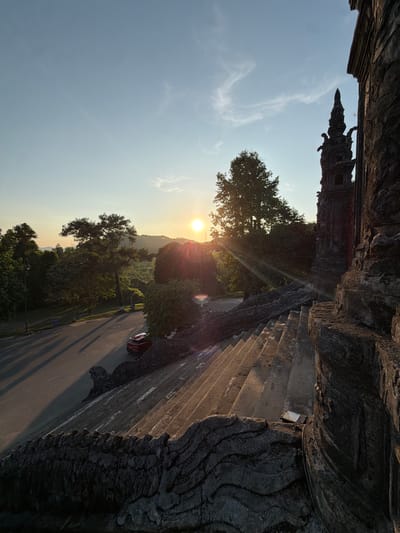 The image depicts a view in Thủy Xuân Ward, Vietnam, captured at dusk or dawn. A multi-tiered tower stands silhouetted against the sky in the upper left. The sun, appearing low on the horizon, is partially obscured by a tree, casting rays across the scene. A road runs through the frame, with a single car visible. The right side of the image is dominated by a textured, wall-like structure, possibly part of a building or monument. The foreground features dense foliage.