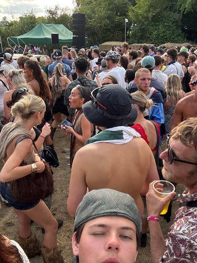 A densely packed crowd fills an outdoor venue. A partly nude person with red underwear and green-and-white cloth draped on their backside is visible. A man wearing sunglasses and holding a drink looks downwards. Another person wearing a tweed cap is facing the camera. A stage with a tent can be seen in the background, along with trees. Several individuals are seated or squatting on the ground within the crowd.