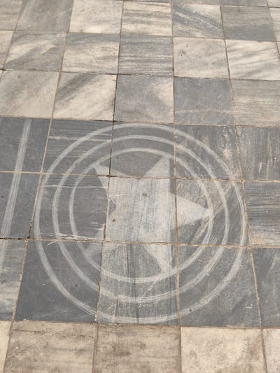 The image shows a tiled stone surface with a faint circular design containing a five-pointed star at the center. The tiles are gray and off-white, arranged in a grid pattern. The circular design, consisting of three concentric rings around the star, is lighter in color, as if it has been partially worn away or cleaned. The location is unknown.