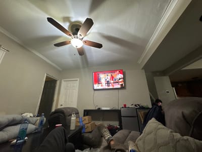 This image depicts an interior living room, identified as being in South Bend, United States. The room features light-colored walls with white crown molding and a white ceiling from which a five-blade brown ceiling fan with a central light fixture is suspended and illuminated, casting shadows. A flat-screen television is mounted on the far wall, displaying a scene with multiple individuals in colorful clothing.
Below the television, a dark console table supports an unidentified electronic device and is situated next to a silver/black compact refrigerator. Two brown cardboard boxes are present on the floor beside the console table.
In the foreground, a dark-colored coffee table or ottoman holds three plastic water bottles (at least two with blue labels), one white insulated tumbler, and a red plastic cup. Two sets of human legs are visible in the immediate foreground: one set to the left, wearing black pants and white socks, with feet resting on the table; and another set to the right, wearing light-colored pants and shoes with dark soles, with one foot propped.
Several upholstered sofas or recliners, in dark grey or brown, are visible. A person is seated on a sofa in the midground to the right, largely obscured by furniture and draped with a dark blanket or jacket, with their head partially visible. Another sofa to the left is covered with blankets or cushions. In the background, two doors are present: a partially open dark door to the left, and a closed white door to the right.