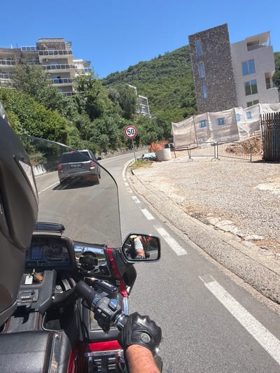 The image depicts a point-of-view shot from a motorcycle traveling on a paved road in Podličak, Montenegro. The road curves to the right, with a steep, tree-covered hillside on the left and a construction site on the right. A silver SUV with license plate AB 799 JU is ahead on the road. A circular speed limit sign indicating "50" is visible on the right side of the road. Modern apartment buildings are under construction on the hillside and adjacent to the construction site. The motorcycle has a red and black color scheme, with a visible dashboard and handlebars. The rider is wearing black motorcycle gloves. The sky is clear and blue.