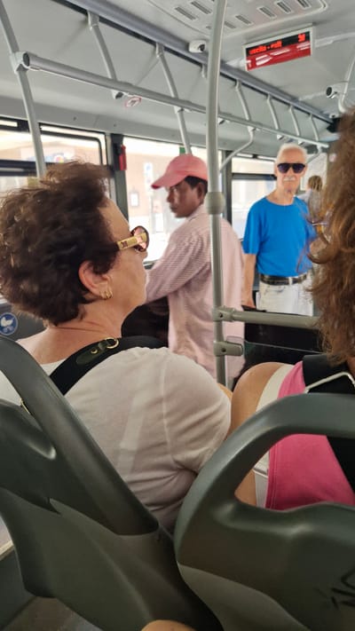 The image shows the interior of a bus in Taranto, Italy. Several passengers are visible, including a man in a blue t-shirt and sunglasses, a man wearing a pink baseball cap, and a woman with dark curly hair wearing sunglasses and gold earrings. Another person is partially visible in the background wearing a pink top. The scene appears to be during the daytime, with natural light coming through the windows. Some notable details include the bus's interior fixtures, such as overhead handrails and seating. A red sign with white text is visible on the left side of the image, possibly indicating an exit. The overall mood seems casual, with people appearing relaxed on their journey.