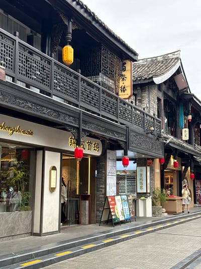 The image shows a street scene in Qingyang District, China, featuring traditional Chinese architecture. The buildings have dark wooden facades with intricate latticework on balconies and decorative carvings. Red lanterns hang from the eaves, and yellow lanterns are also visible. The street is paved with stone, and there are yellow tactile paving strips along the edge of the sidewalk.

On the left side of the street, a storefront displays bamboo plants and mannequins through a large window. Above the entrance, a golden sign reads "shengshouain" in English and Chinese characters. Next to it, another storefront has a golden sign with Chinese characters and a red lantern hanging nearby.

Further down the street, a woman in a yellow jacket and patterned skirt stands near a shop entrance, talking on her phone. Several A-frame signs with colorful graphics and text are displayed on the sidewalk, advertising products or services. The sky is overcast.