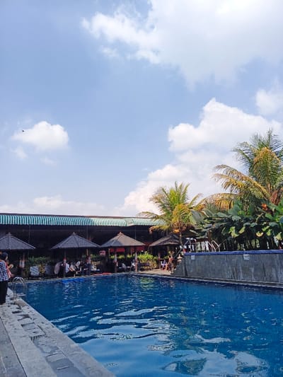 A serene swimming pool dominates the foreground, reflecting the blue sky dotted with fluffy white clouds. Four gazebo-like structures with dark roofs line the pool's edge, providing shaded seating. Palm trees and lush greenery add a tropical feel. A building with a striped green and white awning sits behind the gazebos. Several individuals relax under the shaded structures. The scene exudes a peaceful and inviting atmosphere.