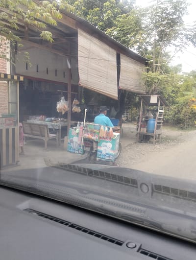 The image depicts a roadside scene in Teupinreudeup, Indonesia. In the foreground, the dashboard and windshield of a vehicle are visible, indicating the photo was taken from inside a car. Beyond the windshield, an open-air structure resembling a shop or market stall is present. This structure has a corrugated metal roof supported by wooden beams, and bamboo blinds are rolled up at the front. Inside the stall, shelves are stocked with various goods, and a table is set up with items on display, including what appear to be snacks in plastic bags hanging from the ceiling. A wooden bench is situated in front of the table.
To the right of the stall, a person wearing a blue shirt and a black cap is operating a three-wheeled cart designed for selling ice cream. The cart is decorated with images of ice cream cones and the word "ES KRIM" (ice cream in Indonesian) and "RASA" (flavor in Indonesian). The cart has a cooler box and other items for sale. Further to the right, a small wooden shelf unit stands independently, holding a blue water barrel and other assorted items.
The ground is covered with gravel and dirt. Lush green trees and vegetation surround the area, suggesting a rural or semi-rural setting within the town. The overall impression is of a typical local business operating alongside a road.
