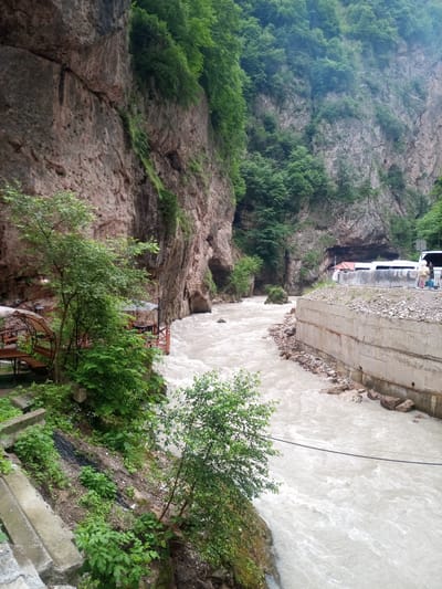 The image depicts a wide, fast-flowing river carving through a narrow gorge in Khushto-Syrt, Russia. The river's water is milky-white, indicating a high sediment load, and churns with visible rapids. Sheer, steep rock faces covered in lush green vegetation rise on both sides of the gorge. On the left bank, a rustic wooden deck with tables and seating is visible, suggesting a potential rest stop or eatery. A few trees and dense green foliage grow along the riverbank and up the rocky slopes. On the right bank, a concrete retaining wall runs along the river, topped with gravel and rocks. Beyond the wall, a paved area is visible with two white buses parked side-by-side and several people standing near them. The sky is overcast, contributing to the muted lighting of the scene.