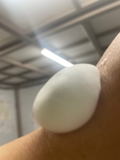 A close-up shot shows a white blob of lotion or cream on a person's skin. The background is out of focus, showing a ceiling with fluorescent lights and a criss-cross pattern of beams, possibly in a warehouse or industrial setting in Tervolovo, Russia. The focus is entirely on the skin and the white substance.
