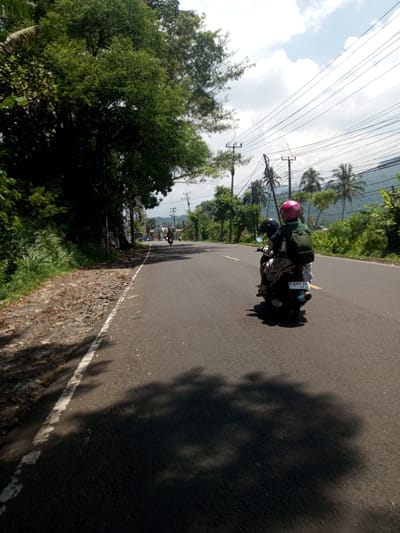 The image shows a paved road in a rural area, with lush green vegetation on either side. A motorcycle with two riders is traveling along the road. The rider in the back is wearing a pink helmet and green backpack. The motorcycle's license plate reads "D 6366 TS". Overhead are power lines and utility poles. In the distance, other vehicles are on the road. The scene suggests a tropical or subtropical climate. There is no evidence that it is in Nawng Hkar, Myanmar.
