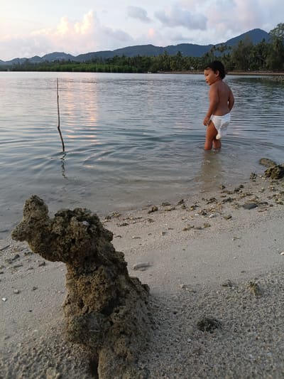 A child is standing in shallow water at a beach in Baan Wok Tum, Thailand. The child, wearing a diaper, is looking down at the water. The water is calm with small ripples. The shoreline is composed of sand and rocks. In the background, there is a mountain covered in vegetation. The sky is cloudy.