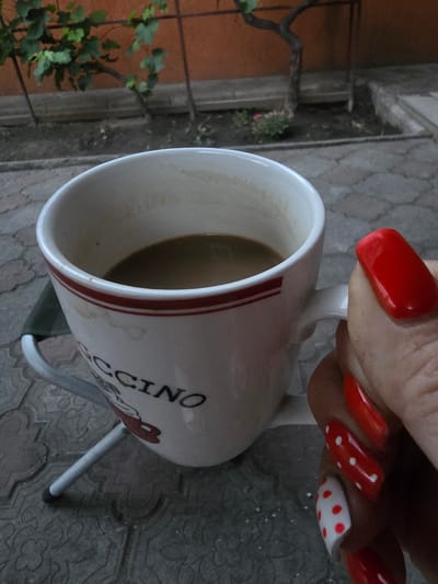 A person's hand with bright red, well-maintained nails is holding a white ceramic mug. The mug is partially filled with a light brown liquid, likely coffee. The mug has a design on it, featuring red stripes near the rim and the word "CCINO" visible in black lettering. There is also a red heart graphic with a bow on the mug.

The setting appears to be outdoors, with a tiled or paver surface visible beneath the mug and hand. In the background, there is a plant with green leaves and a reddish-brown wall, suggesting a garden or patio area. The lighting indicates it is daytime, possibly overcast or in shade, as there are no strong shadows. The overall mood is casual and relaxed. The location context is Nova Kakhovka, Ukraine.
