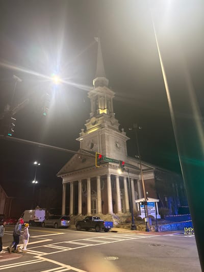 At night, an angled view captures a brightly lit church. The structure boasts a steeple, columns, and detailed architecture. A street sign reads "Lucke St." Traffic lights display red. Cars and pedestrians are visible on the adjacent street, marked with white crosswalk stripes. A lamppost emits a strong glare near the church.