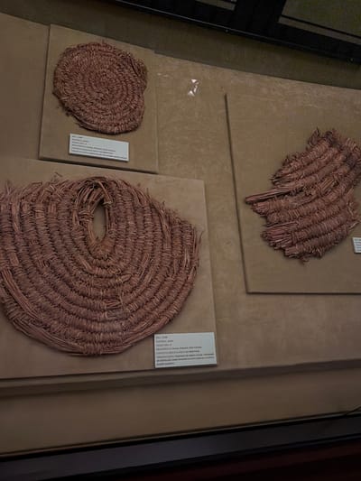 The image shows a museum display case featuring three woven artifacts made of dried plant material, likely reeds or grasses. The display case is divided into sections. On the left, there are two artifacts. The upper artifact is a circular, coiled mat with a textured surface. Below it, a larger, oval-shaped artifact with a central opening is prominently displayed. Its weave is more open and visibly constructed from bundled plant fibers. To the right, a third artifact, a fragmented piece of woven material, is displayed against a textured background. White descriptive labels are visible in front of the two artifacts on the left. The overall environment appears to be a museum with neutral-toned walls and professional lighting. No people are visible in the frame. The artifacts are likely part of an archaeological or ethnographic exhibition in Las Palmas de Gran Canaria, Spain.