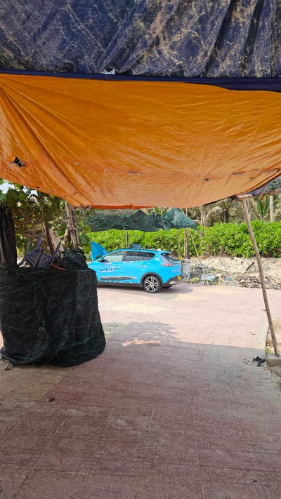 The image shows an outdoor scene in Bac Cam Ranh, Vietnam. A bright blue car with the word "LADO" on its side is parked on a tiled walkway. The area is partially covered by orange and dark blue tarpaulins, and netting is draped nearby. Vegetation is visible in the background. The sidewalk has patterned tiles with engraved circles and lines.