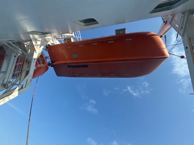 An orange lifeboat is suspended from a white davit against a clear blue sky with scattered white clouds. The lifeboat is viewed from below and to the side, showing its curved hull and a few details on its surface. A thick rope extends downwards from the lifeboat. Part of a ship's white hull and structural elements are visible in the upper left portion of the image. The scene appears to be at sea or in a port. There are no people visible. The city of Unknown, Lithuania is not depicted in this image.
