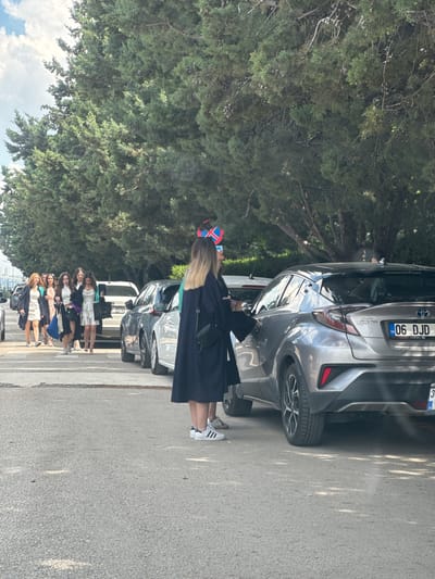 A group of young women, dressed in graduation gowns and academic attire, are seen walking in a line. They appear to be on a street in Etimesgut, Turkey, with a row of trees lining the side of the road and a cloudy sky above. Several cars are parked on the side of the street, and some of the women are interacting with a silver Toyota C-HR, with one woman opening the passenger door. The scene suggests a graduation day celebration, with students preparing to depart after their ceremony. The cars, street, and trees indicate an urban or suburban setting within Etimesgut.