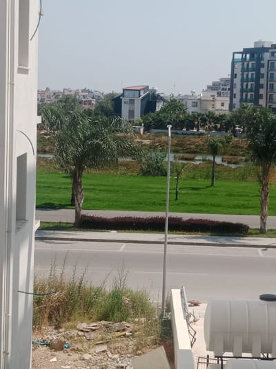 The image depicts an outdoor scene with a view of residential buildings and a roadside area. In the foreground, a white building with a light blue pipe running along its side is visible on the left. The ground is uneven and covered with debris, including rocks and broken tiles, with some dry vegetation.

A paved road runs horizontally across the middle of the image, with a thin strip of dark red flowering bushes bordering the grass on the other side. Beyond the road, a well-maintained grassy area with several palm trees and a small body of water can be seen.

In the background, a cluster of modern apartment buildings is visible. One tall building on the right has a dark facade with multiple floors featuring balconies with teal railings. Further to the left, a lower-profile building with a distinctive angled roof and glass sections is present, along with other residential structures. The sky is clear and light-colored, suggesting a daytime scene under bright sunlight.

The location is identified as Trikomo, Northern Cyprus / Cyprus.