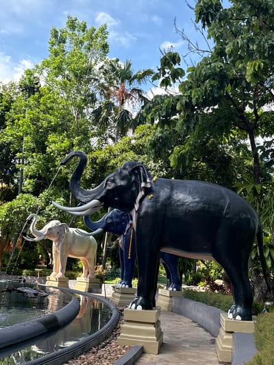 The image depicts a collection of elephant statues in Samrong Nuea Subdistrict Municipality, Thailand. The statues are arranged in a line, with each subsequent elephant smaller than the previous one. The largest elephant is black, followed by a dark blue one, and then two lighter-colored elephants. The statues are positioned on pedestals beside a water feature with a dark, round edge. Lush greenery, including trees with dense foliage, surrounds the statues, with a glimpse of a blue sky and palm trees visible above.