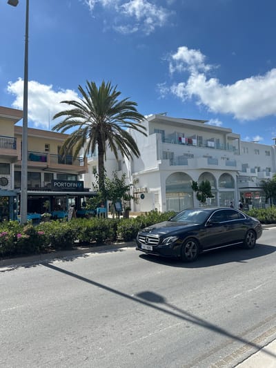 A black Mercedes-Benz E-Class sedan is driving on a paved road. The car has a license plate that reads "TEE 905". In the background, there is a street with a restaurant called "PORTOFINO" which serves "MEDITERRANEAN CUISINE". The restaurant has tables set with blue tablecloths outside. Behind the restaurant is a white multi-story building with balconies. A tall palm tree stands in front of the building. The sky is blue with some white clouds. The scene appears to be in a Mediterranean coastal town, likely in Paphos Municipality, Cyprus, given the style of architecture and vegetation. The overall impression is a bright, sunny day.