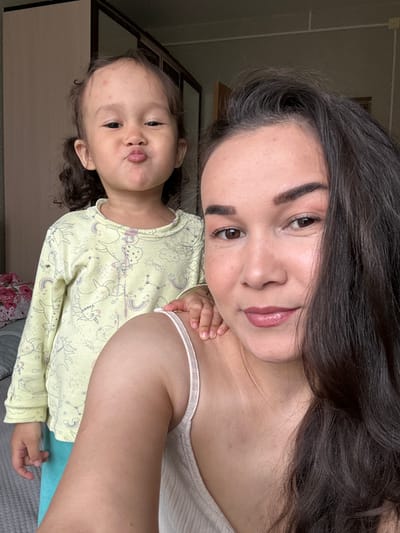 The image captures a selfie-style portrait of a woman and a young child. The woman, with dark brown hair and fair skin, is positioned at the top of the frame, looking down towards the camera. She has dark eyebrows, brown eyes, and is wearing lip gloss. A portion of her shoulder and a thin-strapped tank top are visible.

Below her, the child is wearing a long-sleeved yellow shirt with a unicorn pattern. The child has light skin, brown hair, and is pursing their lips in a pouting expression.

The background shows a muted wall and a partially visible cabinet. The image appears to be taken indoors. The location in the city of Uray, Russia is not discernible from the image.