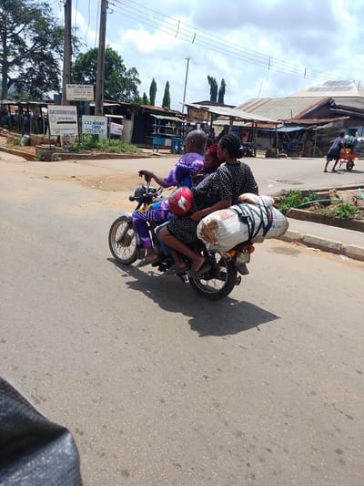 This media file captures a scene on a paved road in Iseyin, Nigeria. The central subject is a motorcycle carrying three people and a substantial load secured to the rear. The driver, wearing a purple and yellow patterned outfit, is at the front. Behind them, a person in a black and white polka dot dress is seated, and a child in a purple garment is cradled in their arms. The load on the back of the motorcycle consists of two large, white sacks, tightly bound with black straps. A red helmet rests on the load.

The background reveals a bustling roadside environment. Several stalls with corrugated metal roofs are visible, suggesting a market or commercial area. Signs are present, including one for "OUR LADY'S CATHOLIC HOSPITAL ATUPA EYE CLINIC," another for "ISLAMIC BROTHERHOOD OF NIGERIA ISEYIN BRANCH," and one for "CSS MOVEMENT CHURCH." The sky is cloudy, indicating a typical daytime with moderate light. The overall atmosphere appears to be that of everyday life, with people engaged in transportation and commerce. The road surface shows signs of wear and tear, typical of many urban and semi-urban areas. The presence of multiple people on a single motorcycle and the large, unwieldy load highlight the practicalities of transportation in this location.