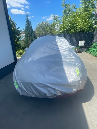 A silver car cover is draped over a car parked on a paved driveway. The car cover is light-colored and reflective, with some visible yellow reflective strips. The driveway is made of interlocking pavers. To the left of the car, a white wall is visible, and to the right, there is a dark fence and some green landscaping elements. In the background, trees and a bright blue sky with scattered clouds are visible. The scene appears to be in a residential area in Мухавецкі сельскі Савет, Belarus, given the context.