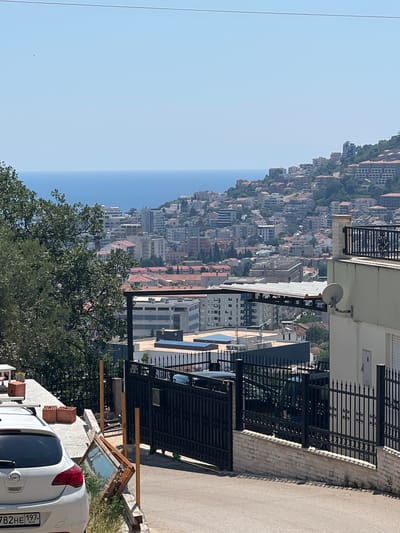 The image captures an elevated view of a coastal city, likely Podostrog, Montenegro, characterized by its dense urban development cascading down a hillside towards the sea. A clear, light blue sky dominates the upper portion of the image, with a sliver of the dark blue sea visible on the horizon. The hillside is densely packed with buildings of various sizes and architectural styles, featuring a predominance of red-tiled roofs alongside modern apartment blocks and hotels.
In the foreground, a white Opel hatchback is parked on a paved surface, with its rear end visible on the left side of the frame. Next to the car, there are two bricks stacked on a concrete ledge. A large black metal fence runs along the edge of the property, separating it from the road below. This fence is supported by a low brick wall. A broken wooden window frame with glass is propped up against a vertical wooden pole next to the fence, leaning towards the ground.
Further down the slope, more urban structures are visible. A prominent multi-story building with a light-colored facade and a black wrought-iron balcony railing stands on the right side of the frame. A satellite dish is attached to the wall of this building. Adjacent to it, a black metal gate is part of the boundary of another property, with a dark-colored vehicle partially visible behind it. A large, flat-roofed modern building with rows of windows is situated in the middle ground, partially obscured by trees.
The overall scene depicts a sunny day in a developed coastal area, with a mix of residential and commercial buildings, and the natural element of the sea in the background. The foreground elements suggest ongoing construction or maintenance work, with the stacked bricks and the discarded window frame.