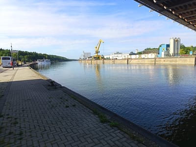 The image depicts a wide canal or river in an industrial port area. On the left bank, a paved walkway with interlocking grey bricks runs alongside the water. A white tour bus is parked on the walkway, and a white passenger boat is docked further down. The right bank of the canal features a concrete quay lined with industrial buildings and infrastructure, including a prominent yellow gantry crane and several silos. The sky is blue with scattered white clouds, and the sun casts reflections on the water. The scene appears to be a calm, sunny day in a working port. This is a view of the Nuremberg port, specifically the Nuremberg harbor basin or an adjacent waterway within the port complex.