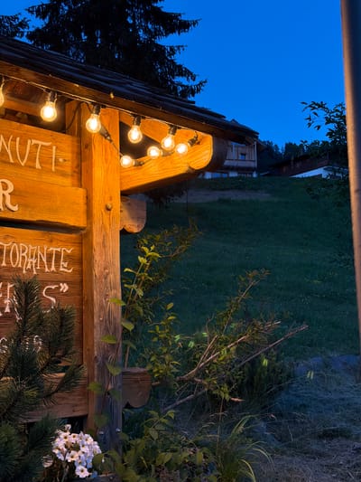 The image depicts an outdoor scene at dusk, likely in or near Lateis / Latais, Italy, focusing on a wooden sign with a string of illuminated bulbs hanging from its overhang. The sign, made of rough-hewn wood, has white lettering that partially reads "NUTI" and "TORANTE". To the left of the sign, a portion of a pine tree and a cluster of white flowers are visible. The background shows a grassy slope with a few rustic buildings nestled into the hillside, silhouetted against a deep blue twilight sky. The lighting suggests an evening ambiance, with the string lights casting a warm glow. The environment appears to be a rural or mountainous area, consistent with the region of Lateis / Latais.