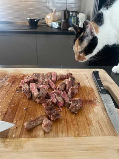 A calico cat is looking at sliced steak on a wooden cutting board. The steak is seasoned with salt and is on the left side of the cutting board, while a knife is on the right. The background shows a kitchen counter with a pot and other items, suggesting the scene is indoors. The cat is partially visible in the upper right corner of the image.