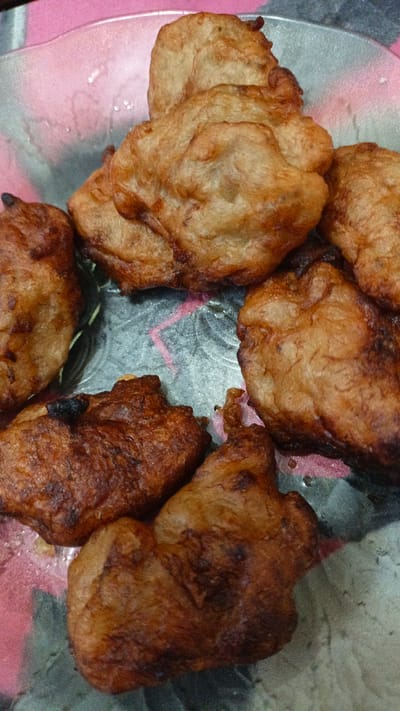 A close-up, overhead shot displays a cluster of deep-fried, golden-brown fritters arranged on a clear glass plate. The fritters, appearing somewhat irregular in shape and texture, suggest they are likely a sweet pastry or savory snack. They are placed on a pink and grey patterned surface beneath the transparent plate, visible through the glass. The lighting appears to be ambient, casting soft shadows and highlighting the crispy exteriors of the food. No people are visible in the image. The location is a domestic setting in Palohlada, Indonesia.