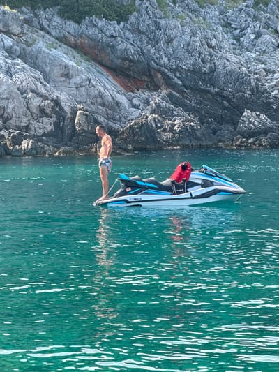 A man is on a jet ski next to a rocky cliff face. The jet ski is blue and white with "YAMAHA" written on it. The man is wearing patterned swim trunks and a red life vest. The water is a clear, turquoise green, and sunlight glints off its surface. The rocky cliff face is gray and textured, with some patches of greenery. The scene appears to be taking place in a coastal area, likely during the day, given the bright sunlight and clear water. The man is positioned on the jet ski, possibly preparing to ride or having just stopped. The overall impression is one of leisure and outdoor recreation in a scenic natural environment.