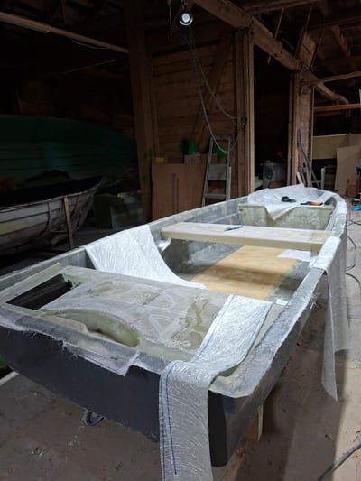 In an indoor workshop, likely in Žemaitkiemis, Lithuania, a fiberglass boat is under construction. The boat, made of a dark grey material, is partially covered with white fiberglass matting and what appears to be resin. The interior of the boat features a wooden plank seat and a smaller compartment. Rolls of fiberglass matting are visible on the edges of the boat, suggesting an ongoing application process. In the background, other boats, including a green one, are stored in the dimly lit space. Wooden structures and tools, such as a ladder and chains, are also present in the workshop environment.