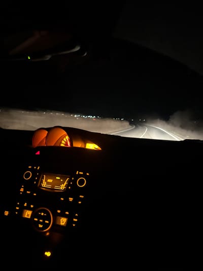 The image displays a nighttime scene from the interior of a vehicle traveling on a road in Padarevo, Bulgaria. The perspective is from the front cabin, looking forward through the windshield. The lower half of the frame is dominated by the vehicle's dashboard, featuring an illuminated infotainment system displaying "AUX" and climate controls showing "23.0" degrees Celsius, all with amber-orange backlighting. Partially visible above the center console are similarly illuminated instrument cluster elements, including a speedometer. Through the windshield, a two-lane paved road is visible, curving to the right. White lane markings are discernible due to the vehicle's headlights. Significant amounts of dust or fine particulate matter are illuminated by the headlights, forming a low-lying haze across the road. In the far distance, a horizontal array of blurry, artificial lights indicates a distant settlement. No individuals are clearly visible.