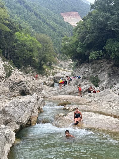 This image captures a scene at the Gola del Furlo Nature Reserve near Cagli, Italy. In the foreground, a young boy swims in a river with a rocky bed and clear, flowing water. Behind him, a young girl sits on a wet, rocky ledge by the river. The surrounding environment is a rugged canyon with steep, rocky walls covered in lush green trees. Further up the canyon, several people are visible on the rocks, some appearing to be resting or preparing to swim. The landscape is dominated by natural rock formations and dense vegetation, with a visible scar from what appears to be a past landslide or quarrying operation on the distant mountainside. The overall atmosphere suggests a recreational outing in a natural, wild setting.