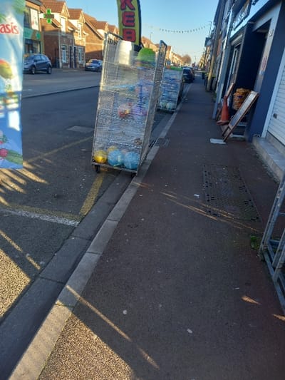 The scene depicts a sunny street in Noyelles-sous-Lens, France, viewed from a slightly elevated perspective towards the north. The primary focus is on two metal roll cages, shrink-wrapped with clear plastic, positioned partially on the curb and pavement of the right-hand sidewalk. These cages contain various colored plastic household items, including bowls and baskets. The front-most cage has its front wheels situated in the street gutter.
To the left, a multi-lane asphalt road runs parallel to the sidewalk, marked with a white line and bordered by a concrete curb. A large, partially visible banner advertising "Oasis" stands on the far left. Shadows from unseen objects extend across the road.
To the right, a paved sidewalk, darker in color than the road, runs alongside a row of commercial buildings. One storefront is dark blue-grey with a white door, featuring a red traffic cone and two A-frame signs placed near its entrance. Several storm drain covers are visible on the sidewalk. A yellow banner with "KEB" partially visible is suspended above the roll cages.
In the background, the street recedes, lined by brick and rendered buildings of varying heights. Parked vehicles are visible on the left side of the street. Overhead, string lights are strung across the street. The sky is clear blue. No individuals are present in the observable frame.