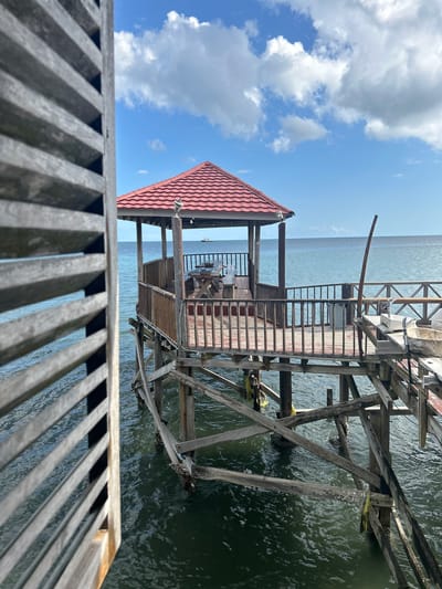 A wooden gazebo with a red tiled roof stands on stilts over the ocean. The gazebo is accessible via a wooden walkway and features wooden railings, a table, and benches. A ship is visible on the horizon. The scene is framed by wooden shutters on the left side of the image. The location is not specified in the city of A R A, Indonesia.