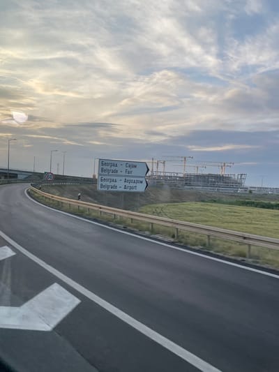 The image shows a view from a car window on a highway leading towards Belgrade, Serbia. Road signs indicate directions to Belgrade Fair and Belgrade Airport. Construction with numerous cranes is visible in the distance. The sky is filled with clouds. A speed limit sign shows 60. The location is likely in Surcin Urban Municipality, Serbia, based on the road signs pointing to Belgrade Airport, which is located in this municipality.